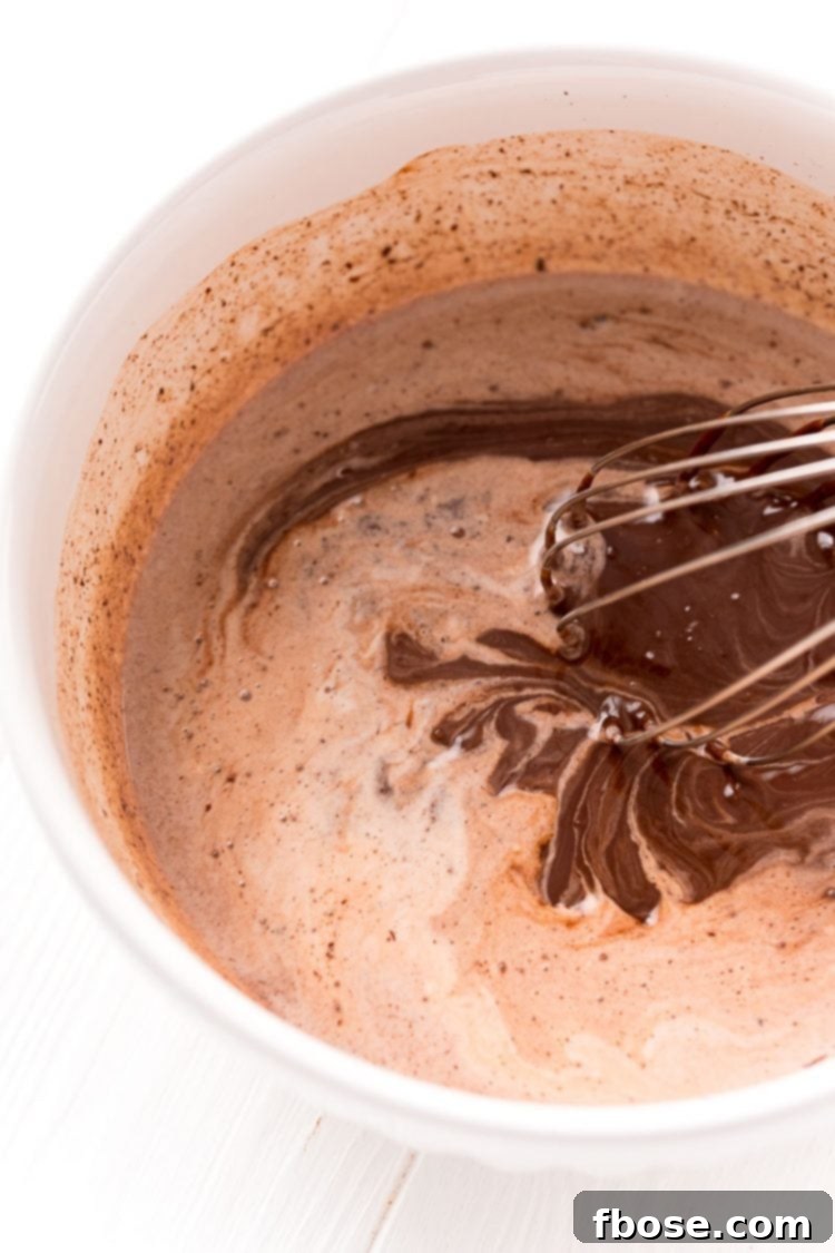 Chocolate and cream being vigorously whisked together in a white bowl, transforming into a smooth, shiny ganache.