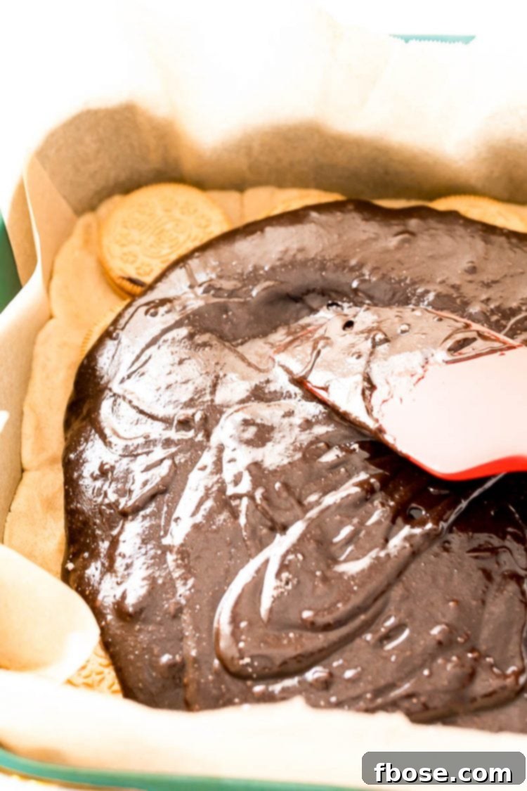 Close-up of a rubber spatula smoothly spreading rich brownie batter over a layer of Oreos and cookie dough in a baking dish.