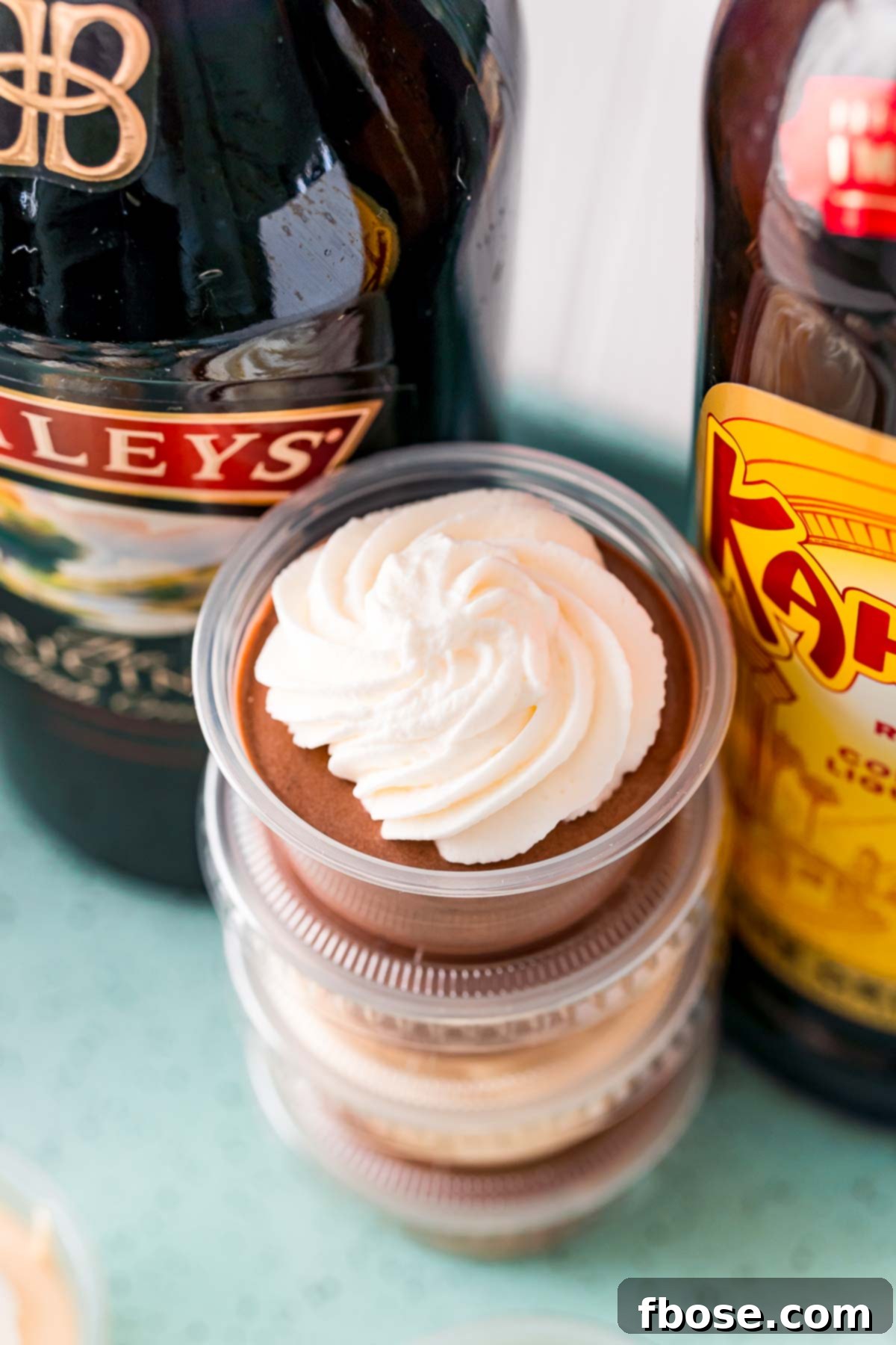 Close-up photo of a stack of creamy, garnished pudding shots positioned between two bottles of liqueur, emphasizing their party-ready appeal.