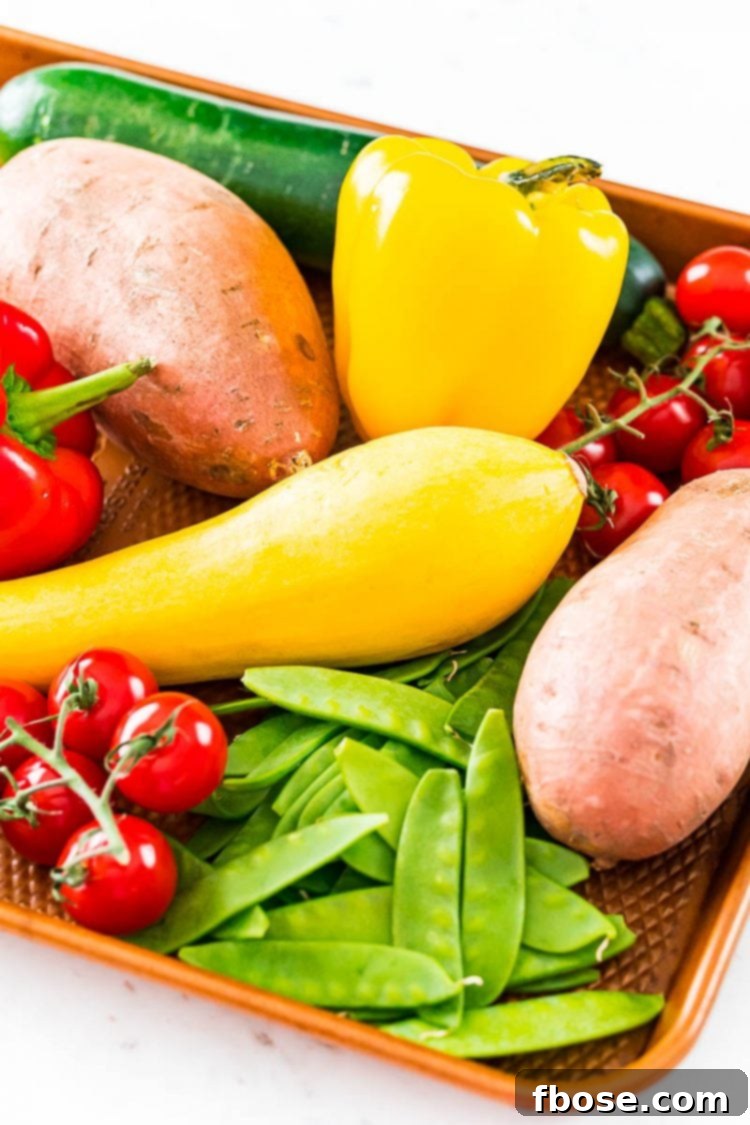A sheet pan loaded with a diverse assortment of fresh, whole vegetables, including bell peppers, zucchini, summer squash, cherry tomatoes, and sweet potatoes, awaiting preparation for roasting.