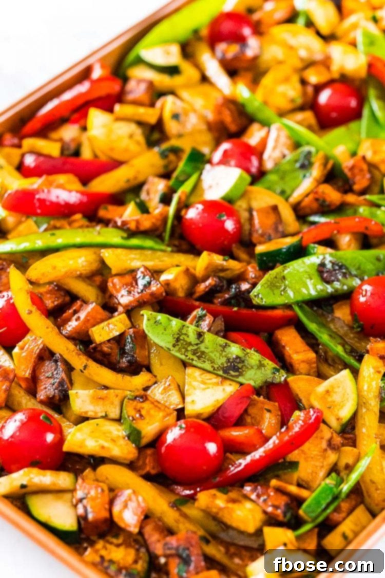 A vibrant assortment of fresh vegetables, including sweet potatoes, bell peppers, zucchini, and cherry tomatoes, coated in dressing and spread in a single layer on a sheet pan, ready for the oven.