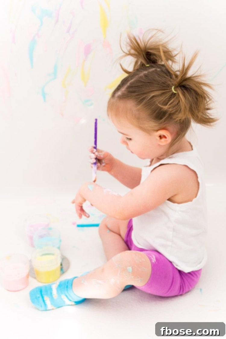 Toddler girl happily painting with bath paint on the bathtub wall.