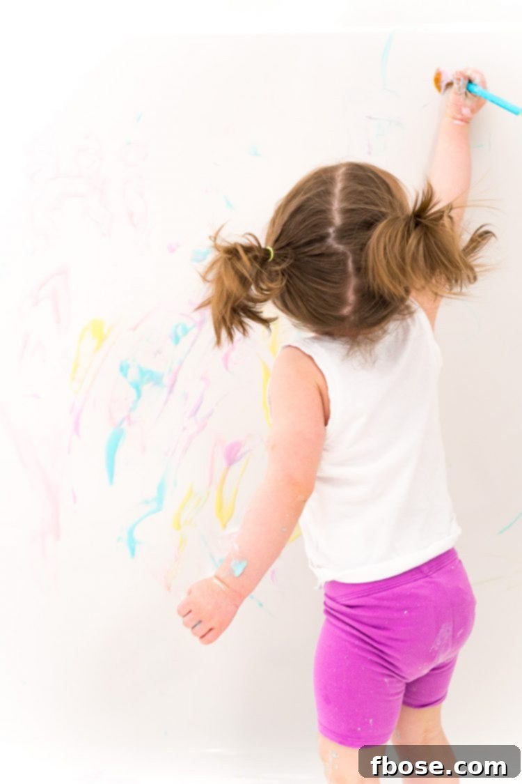 Toddler girl painting the tub with colorful bath paint, creating a vibrant mess.