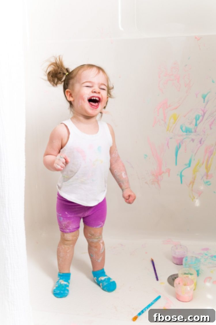 Toddler girl playfully painting with vibrant bath paints in a shallow tub.