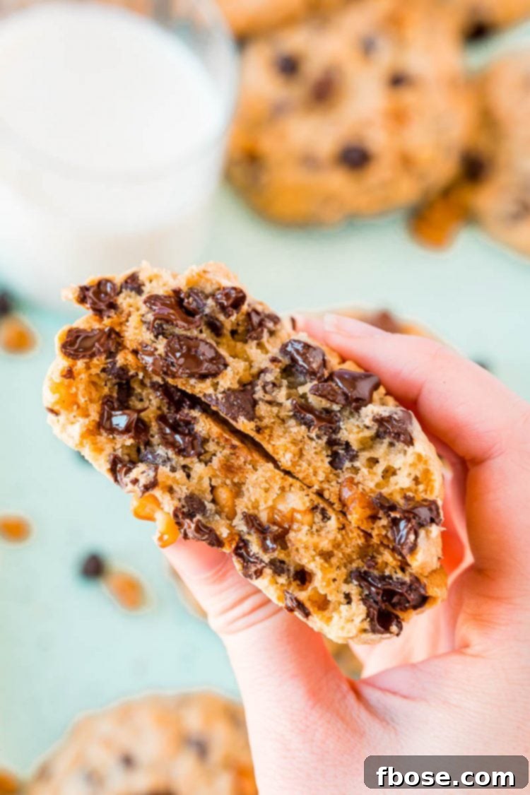 A woman's hand holding a chocolate chip cookie broken in half, revealing its chewy interior and melted chocolate chips.