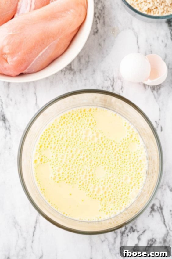Milk and eggs beat together in a glass bowl with raw chicken sitting next to it on a marble countertop.