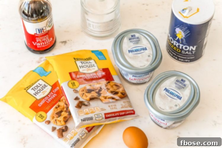 Ingredients for chocolate chip cheesecake bars neatly arranged on a white table: two packages of cookie dough, a block of cream cheese, sugar, an egg, vanilla extract, and salt.