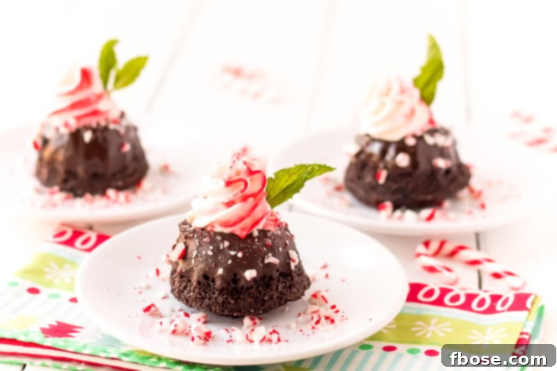 Three Mini Chocolate Peppermint Bundt Cakes arranged on a rustic wooden board, each featuring a dark chocolate base, a glossy ganache drip, and a delicate swirl of pink peppermint whipped cream, adorned with crushed peppermint candy.