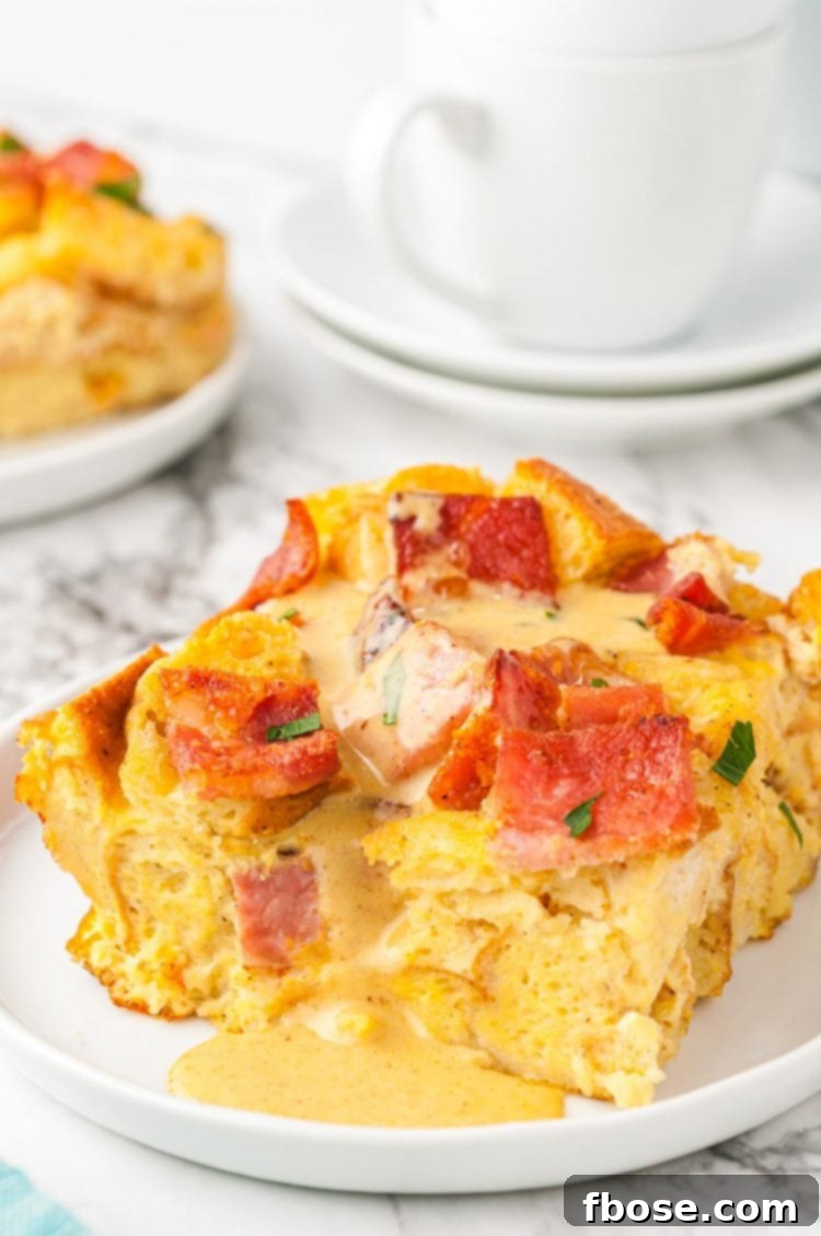 A golden-brown slice of Eggs Benedict Casserole on a white plate, drizzled with creamy Hollandaise sauce.
