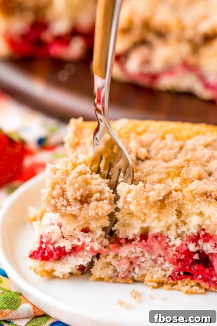 A fork taking a bite out of a strawberry coffee cake slice on a white plate.