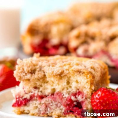 A slice of strawberry coffee cake on a small white plate with a strawberry sitting next to it. More coffee cake in the background.