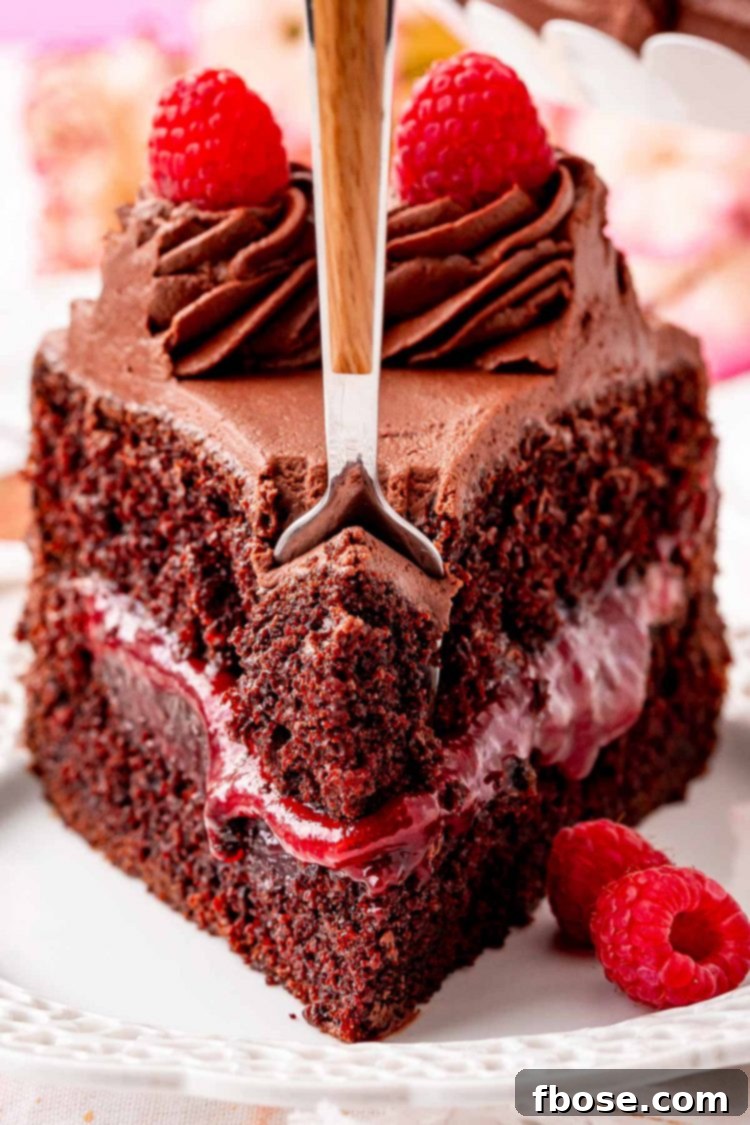 Close up photo of a fork taking a bite out of a slice of chocolate raspberry cake on a white plate.