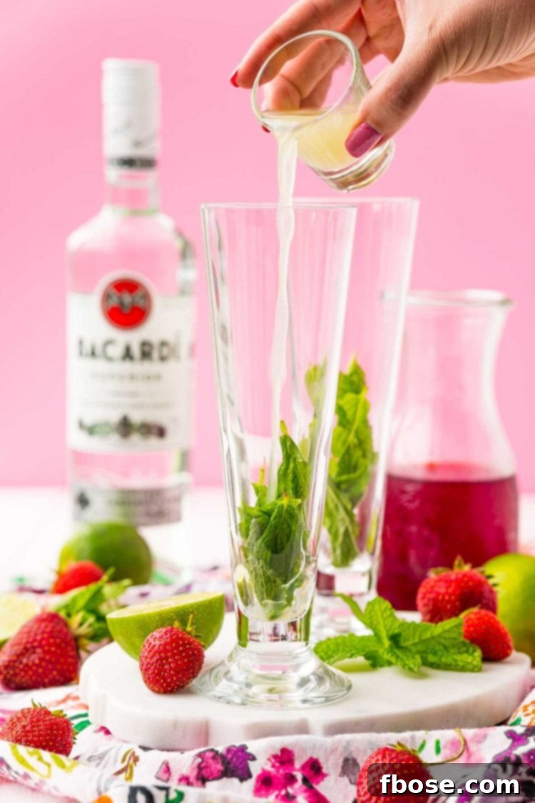 A small shot glass of freshly squeezed lime juice being poured into a tall cocktail glass already containing mint leaves, capturing the vibrant green liquid in motion.