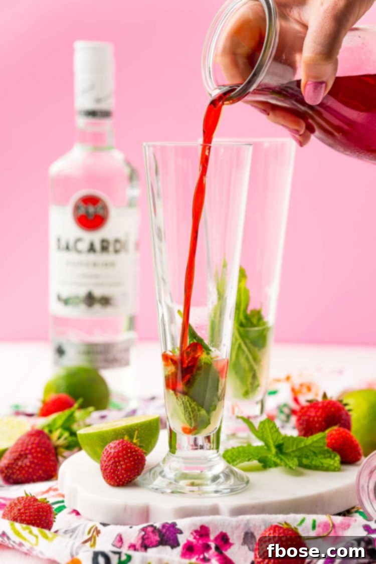 A stream of vibrant red strawberry simple syrup being poured into a tall cocktail glass, joining the lime juice and mint leaves already inside, ready for muddling.