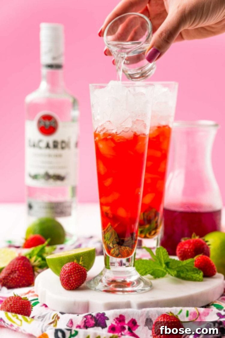 A shot of white rum being poured into a cocktail glass filled with crushed ice, mint, lime, and strawberry syrup, midway through the mojito preparation.