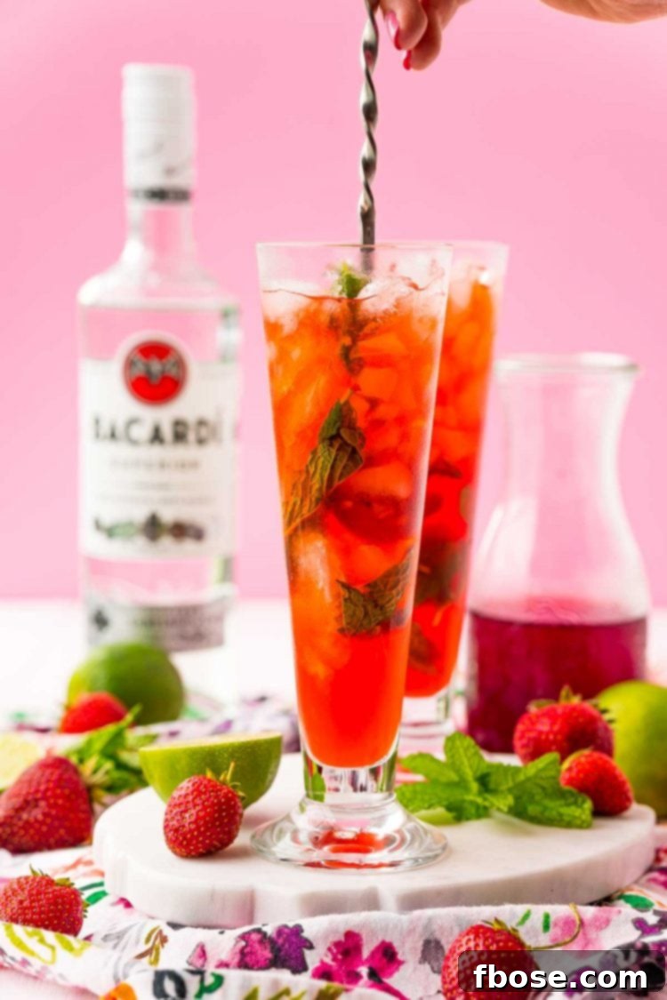 A woman's hand expertly holding a bar spoon, stirring a vibrant red strawberry mojito in a tall glass, illustrating the mixing stage of cocktail making.