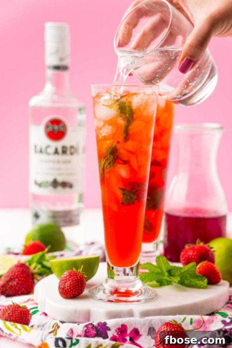 Sparkling club soda being poured from a bottle into a nearly complete strawberry mojito, creating a satisfying fizz and filling the glass to the brim.