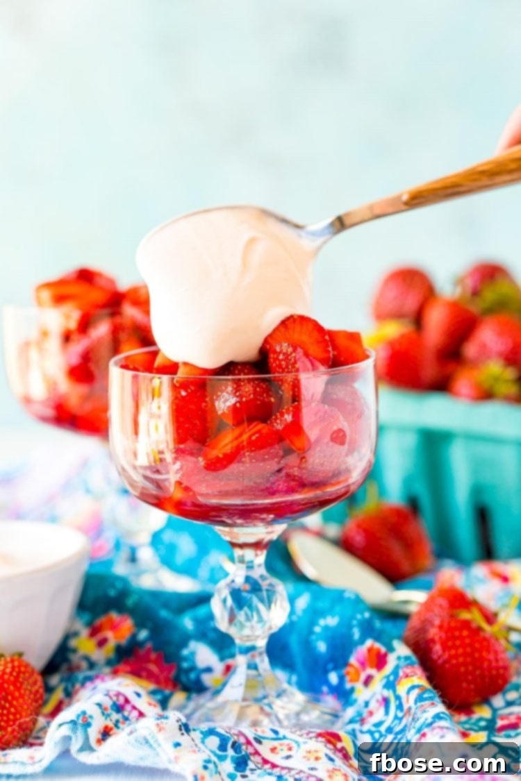 Cream sauce being spoons over macerated strawberries in a stemmed glass to make Strawberries Romanoff.