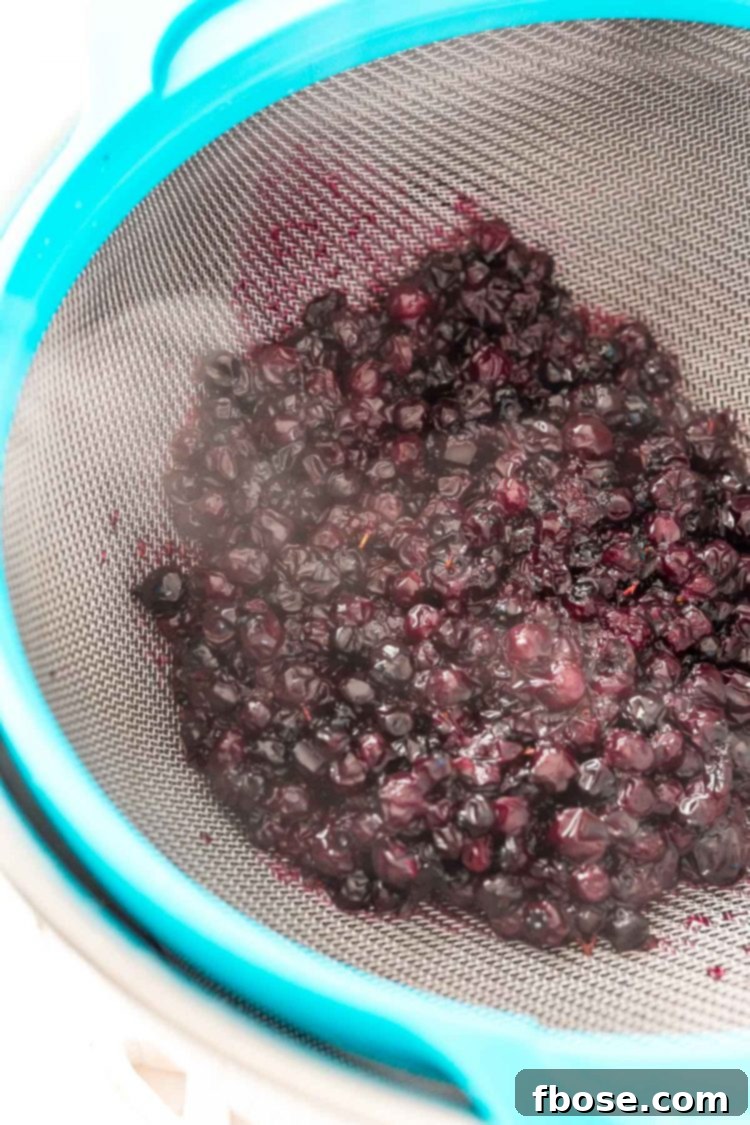 Blueberry Bliss Syrup 5 Cooked blueberries being strained through a fine mesh strainer into a bowl, separating the syrup from the pulp.