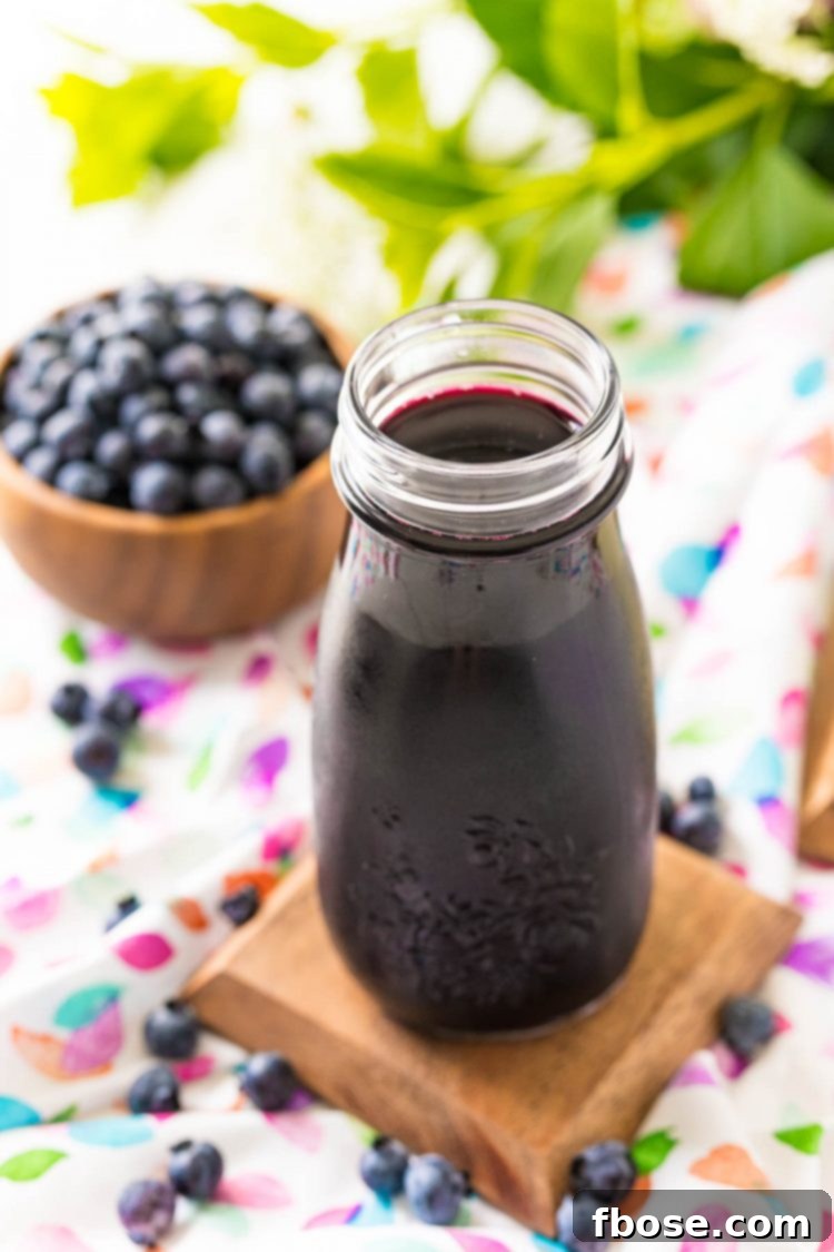 Blueberry Bliss Syrup 6 A small glass bottle filled with clear purple blueberry simple syrup, with fresh blueberries scattered in the background.