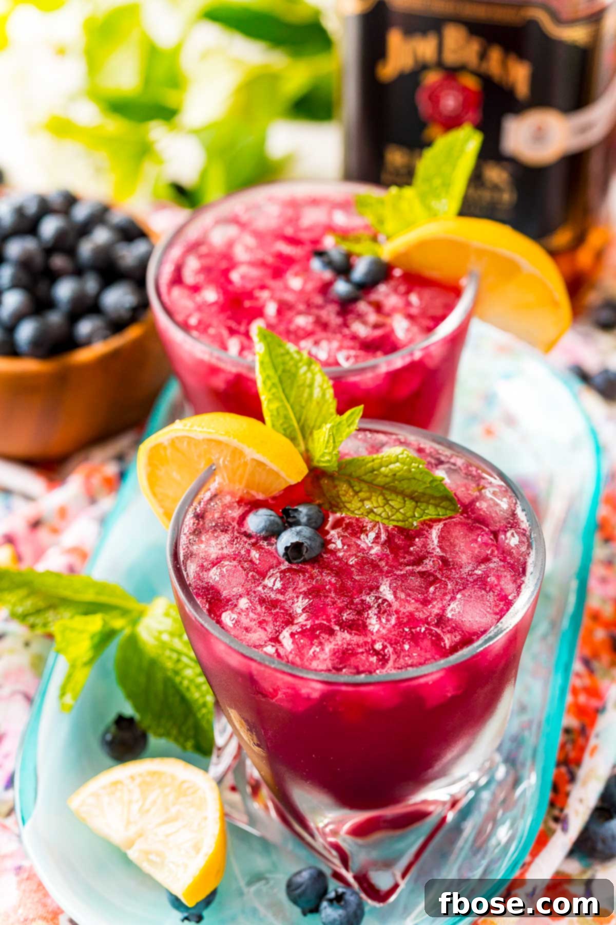 Blueberry bourbon smash on a blue serving tray with lemons, mint, and blueberries scattered around.