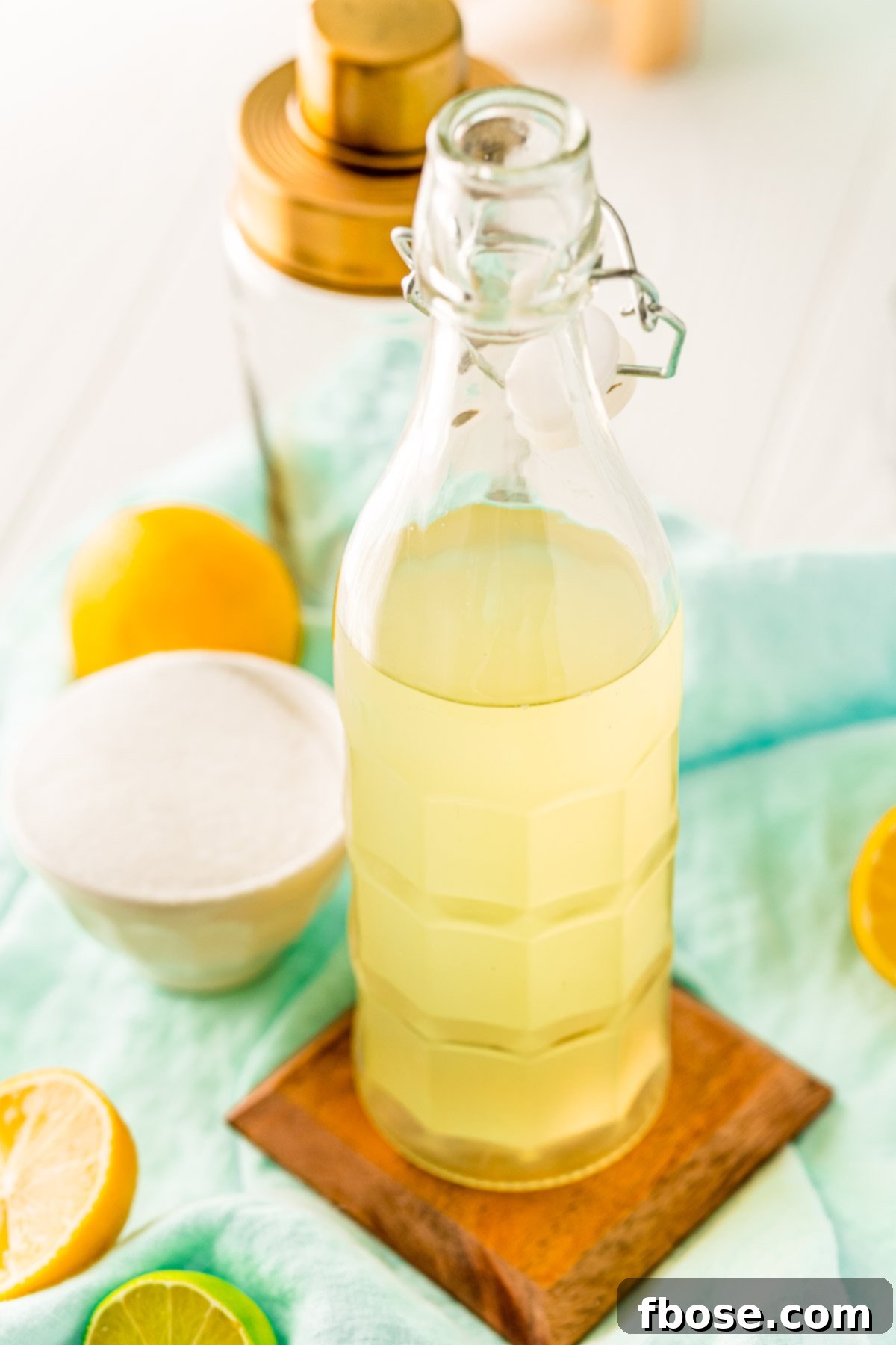 Bottle of homemade sour mix with lemons and limes around it and a cocktail shaker in the background.