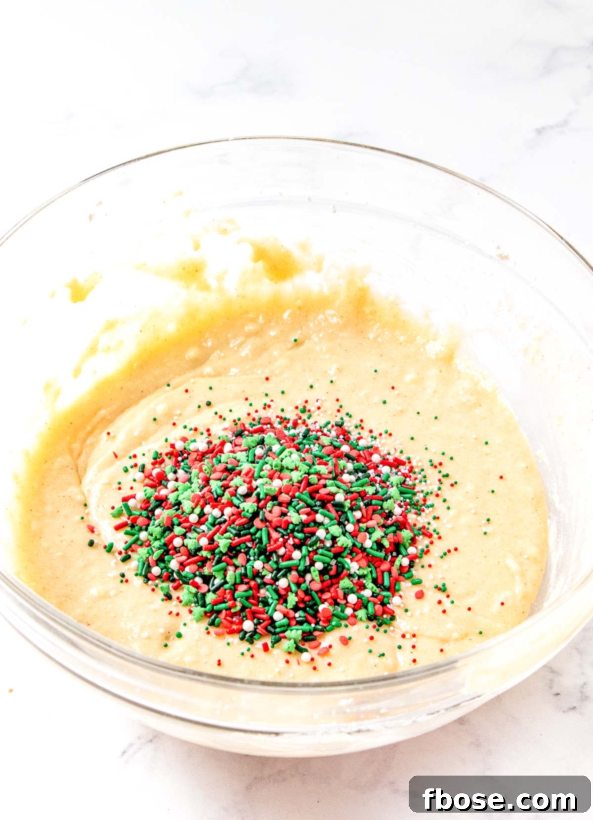 Christmas sprinkles being added to a glass mixing bowl filled with batter for quick bread, demonstrating the final mixing step.