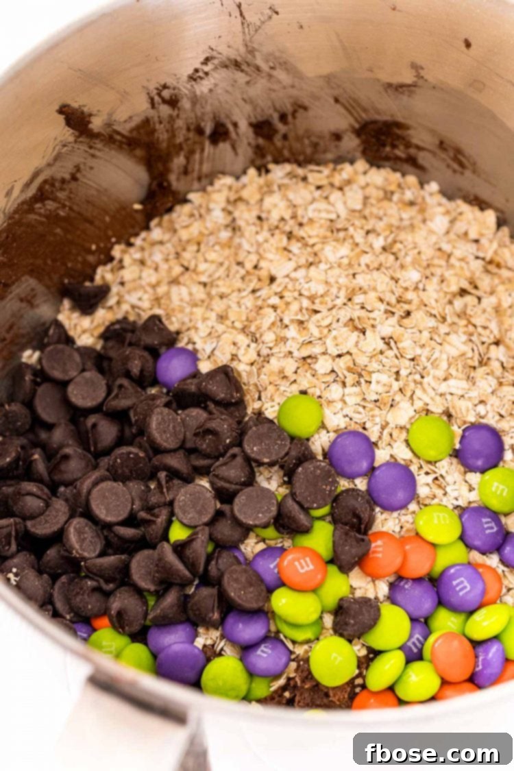 Oatmeal, chocolate chips, and M&Ms being added to a mixing bowl.
