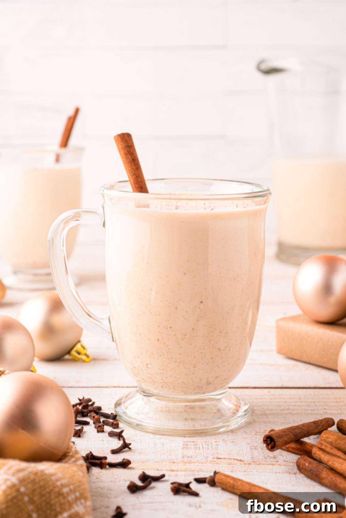 A beautifully styled clear glass mug filled with frothy homemade eggnog, elegantly placed on a table adorned with festive holiday decorations and scattered whole spices, creating a cozy and inviting scene.