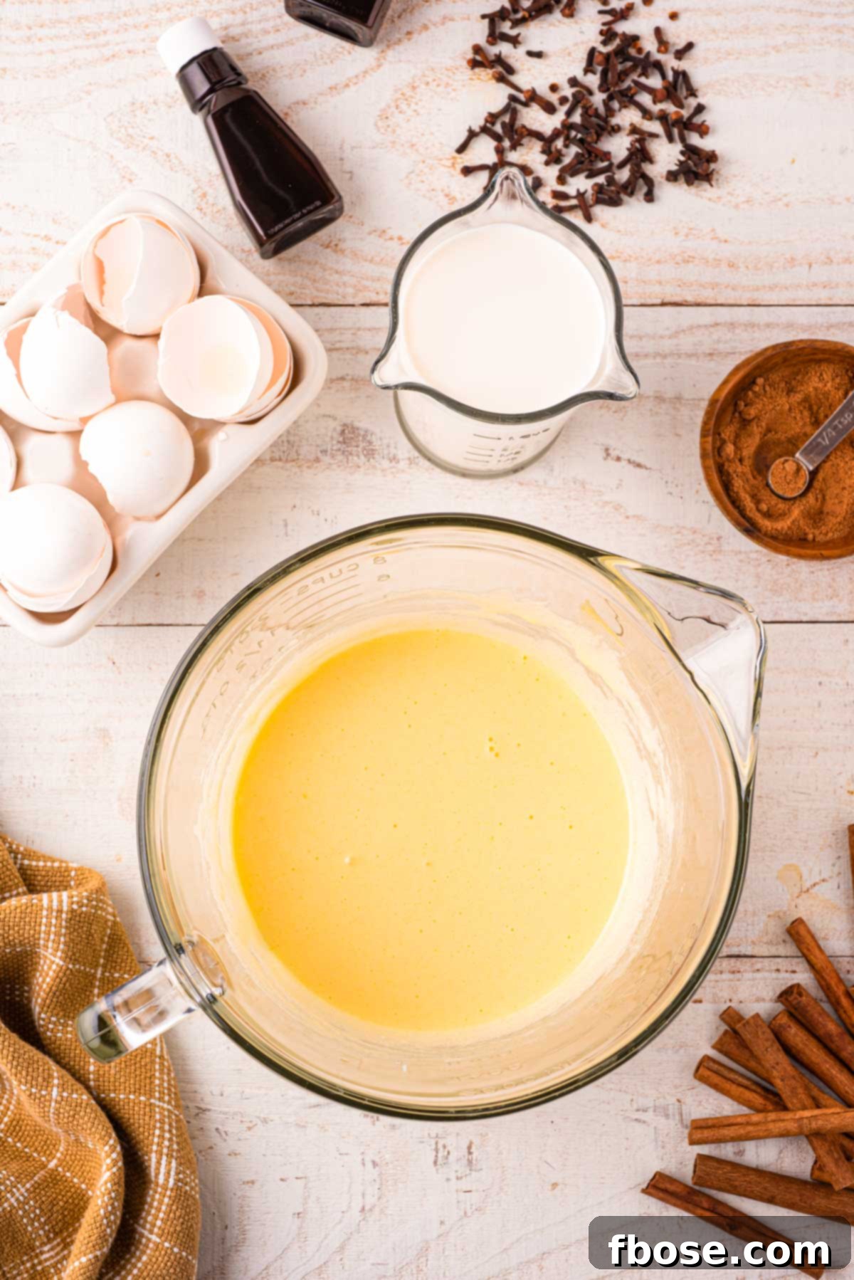 Close-up photo of egg yolks and granulated sugar expertly whisked together to a pale, creamy consistency in a large glass pitcher, signifying the initial step of the eggnog recipe.