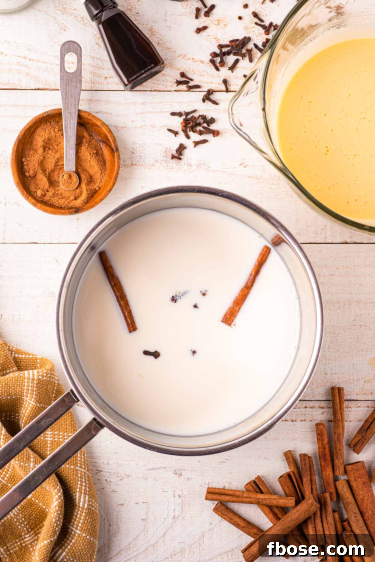 A saucepan on the stove contains milk, cinnamon sticks, and whole cloves, gently heating and infusing the cream with warm, festive spices for the eggnog base.