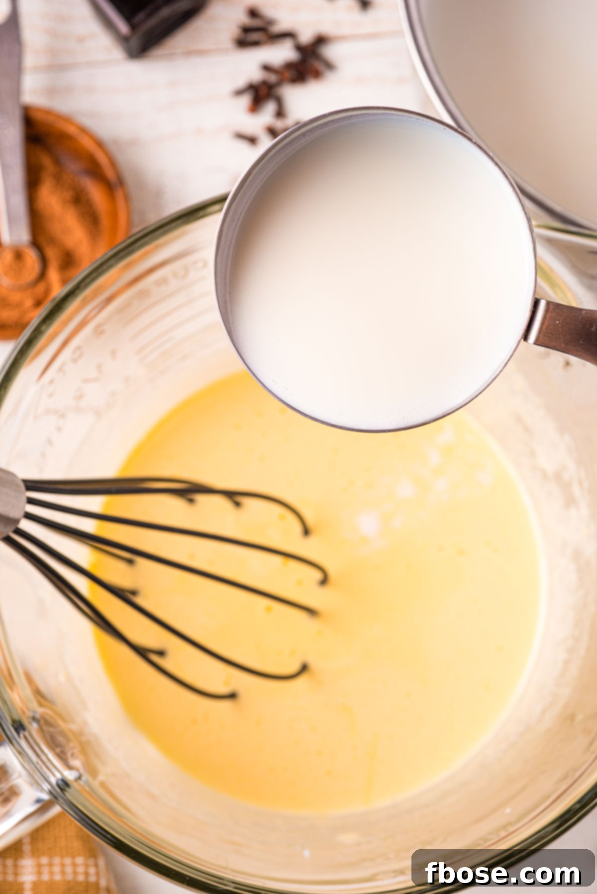 A stream of warm, spiced milk is carefully and gradually poured into a bowl containing the whisked egg and sugar mixture, while a hand continuously whisks to temper the eggs gently.