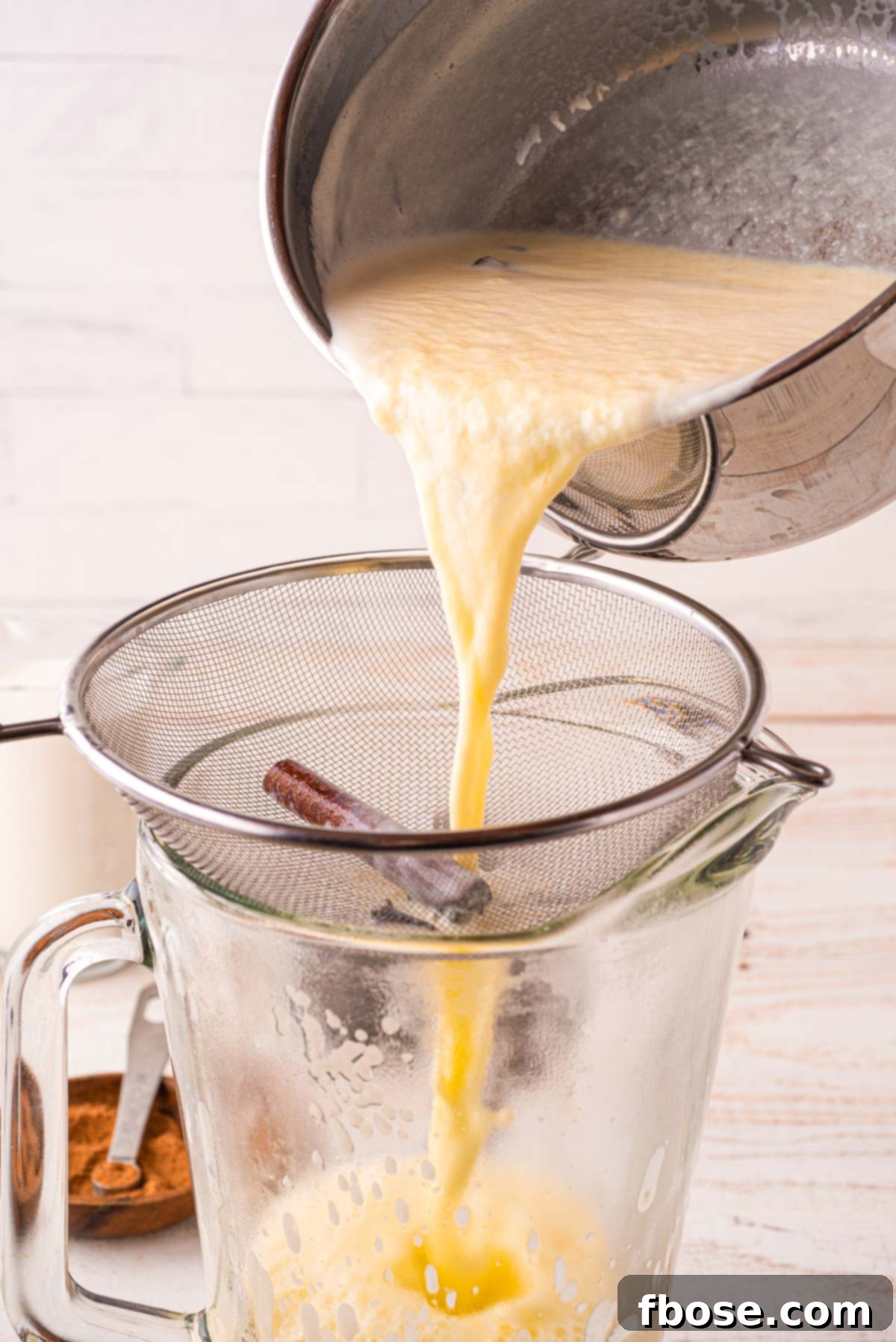 Homemade eggnog is meticulously strained through a fine-mesh sieve into a clear glass jug, ensuring a perfectly smooth and silky texture by removing any solid spices or cooked egg particles.