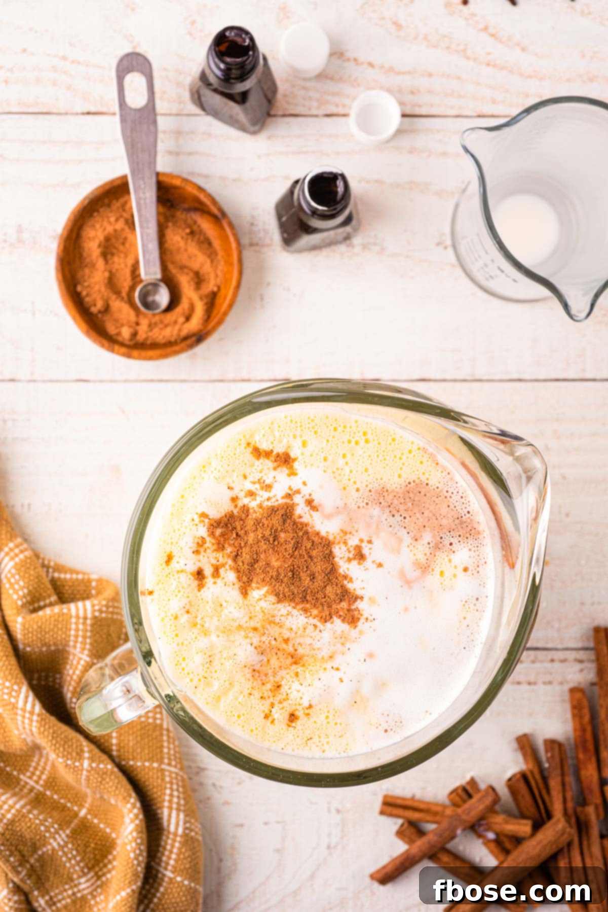 A large glass pitcher filled with the creamy, golden homemade eggnog base, awaiting the final stir-in of vanilla extract, rum extract, and nutmeg, which are delicately resting on the surface.