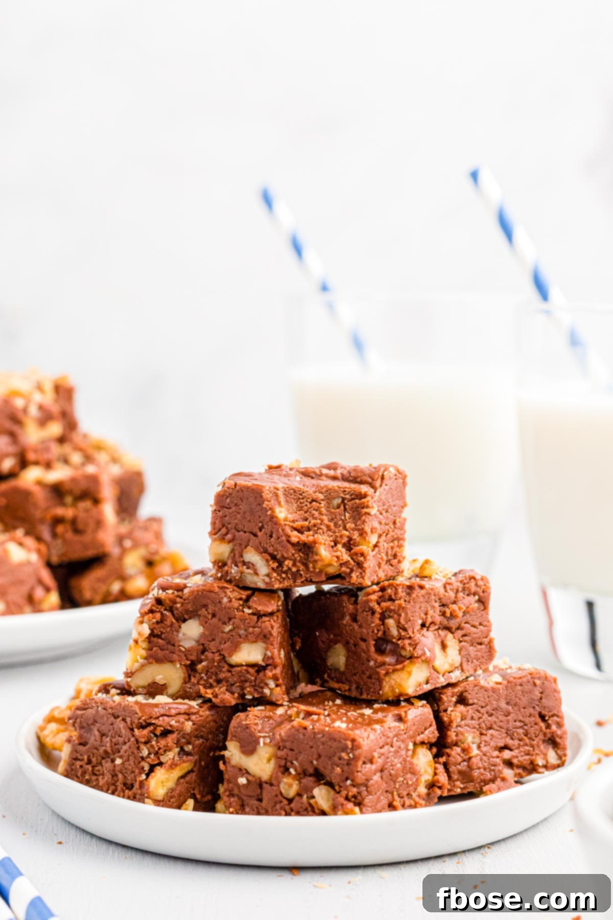 Walnut chocolate fudge squares neatly stacked on a white plate, with blurred glasses of milk in the background, suggesting a delightful pairing.
