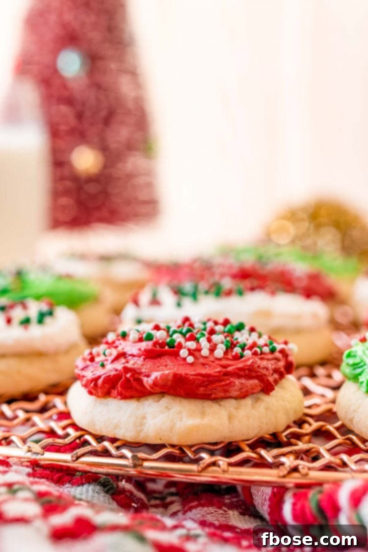 A close-up view of a vibrant red frosted sugar cookie, with additional green and white frosted cookies blurred in the background, showcasing the soft texture.