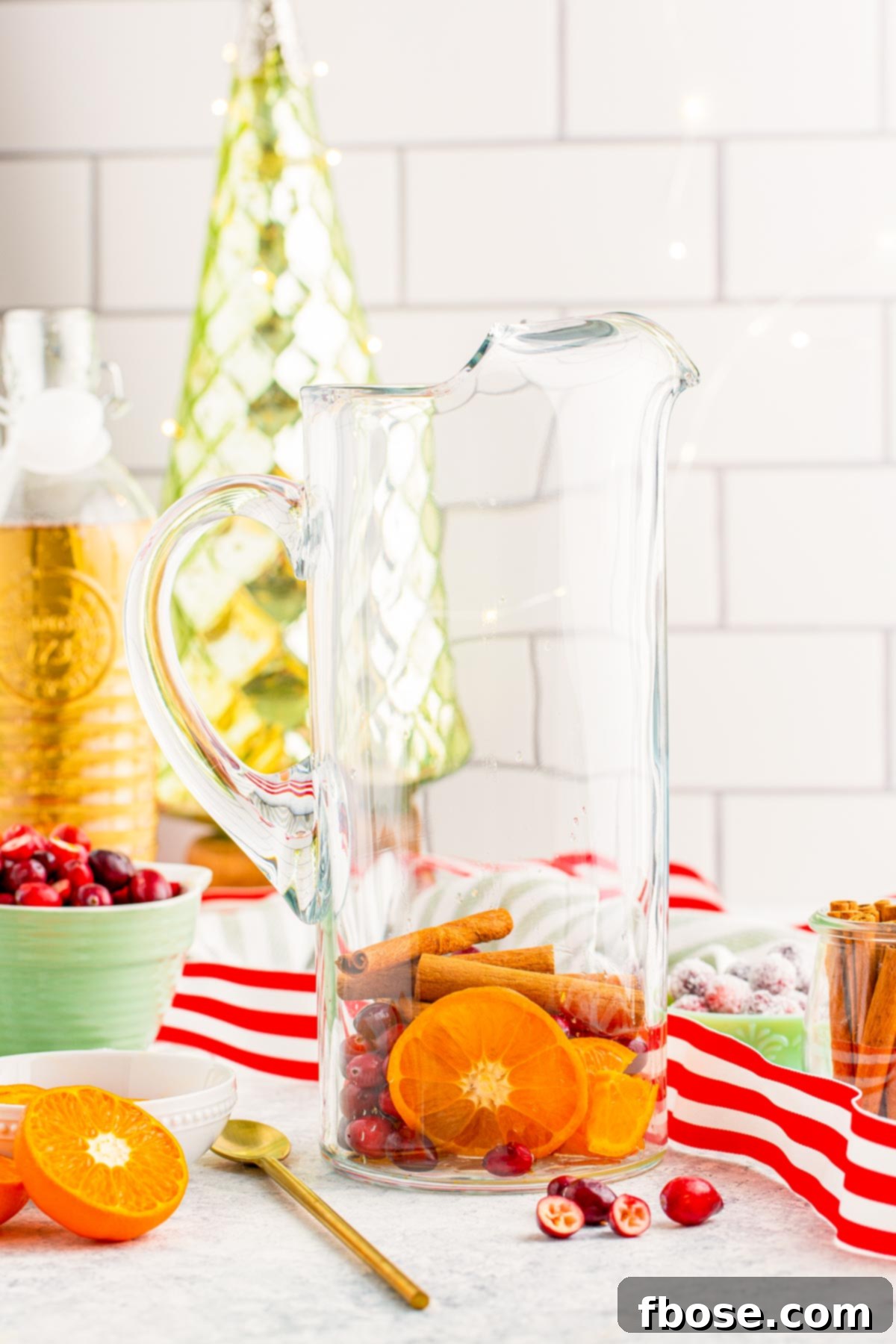 Crimson & Citrus Yuletide Sparkler 6 Fresh orange slices, whole cranberries, and several cinnamon sticks artfully arranged inside a clear glass pitcher, ready for the liquid ingredients to be added for a Christmas punch.