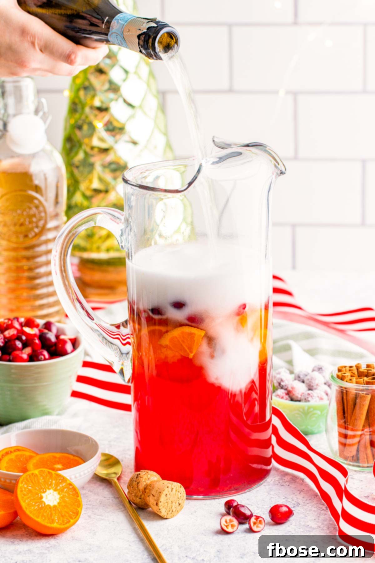 Crimson & Citrus Yuletide Sparkler 7 A bottle of sparkling prosecco being poured into a large glass pitcher already filled with a festive fruit punch, creating a bubbly and inviting Christmas drink.