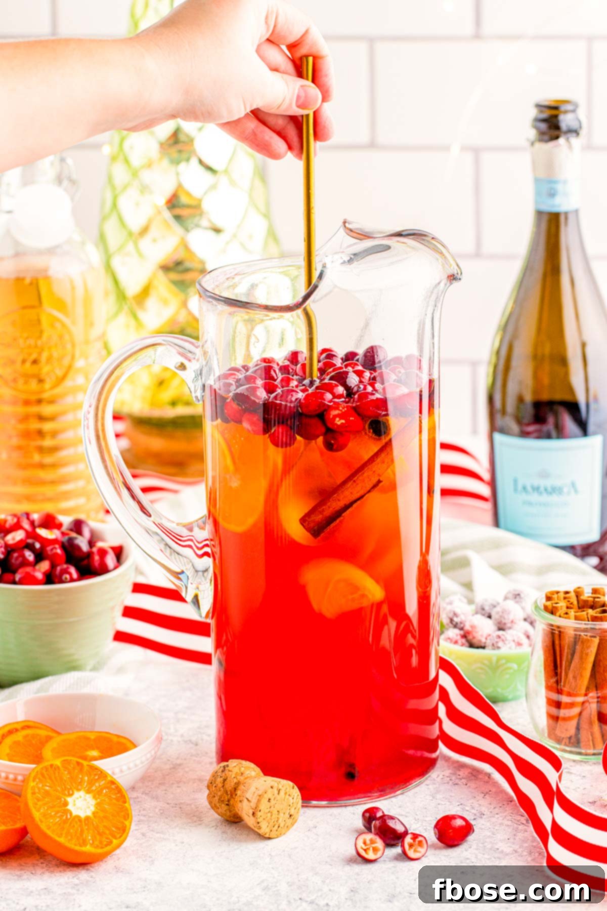 Crimson & Citrus Yuletide Sparkler 8 A woman's hand gently stirring the vibrant Christmas Mimosa Punch in a clear pitcher with a long bar spoon, ensuring all ingredients are well combined and flavors infused.