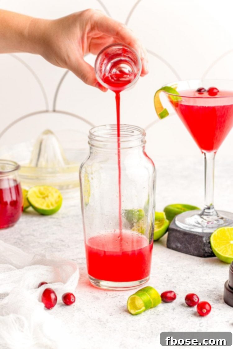 Pouring homemade cranberry simple syrup into a sleek glass cocktail shaker filled with ice and other ingredients, demonstrating the first step in mixing a Cranberry Cosmopolitan.