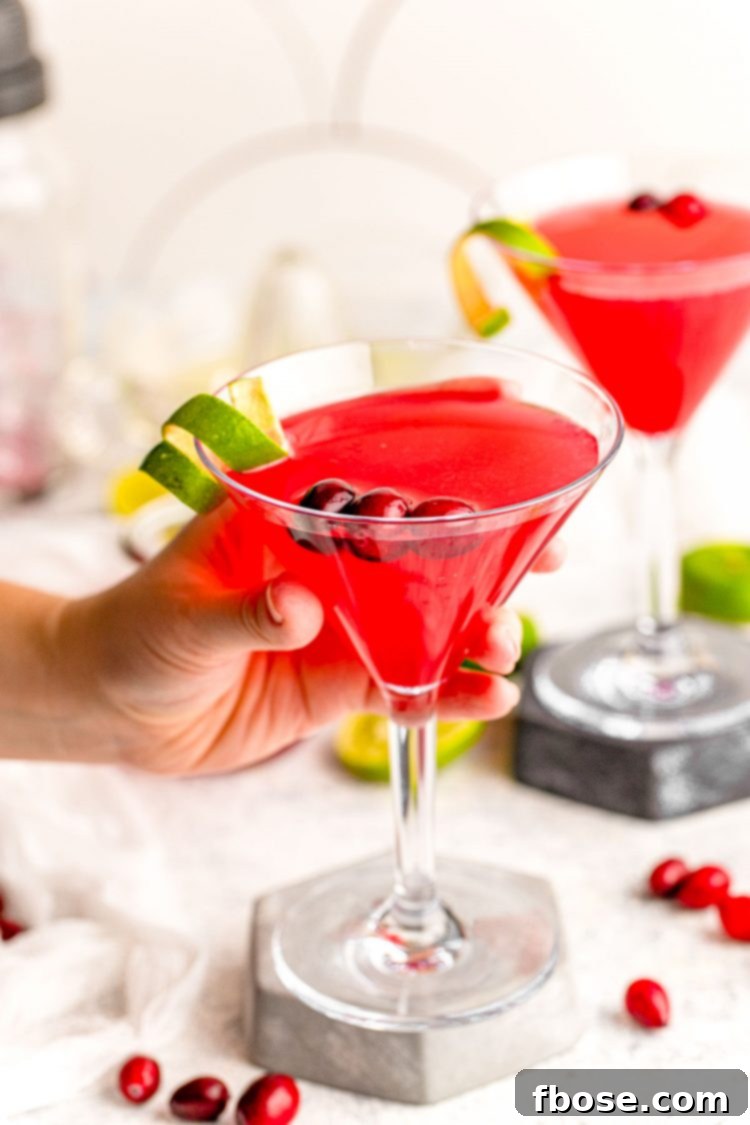 A woman's hand elegantly holding a vibrant Cranberry Cosmopolitan cocktail in a martini glass, set against a soft, inviting backdrop, emphasizing the drink's appeal.