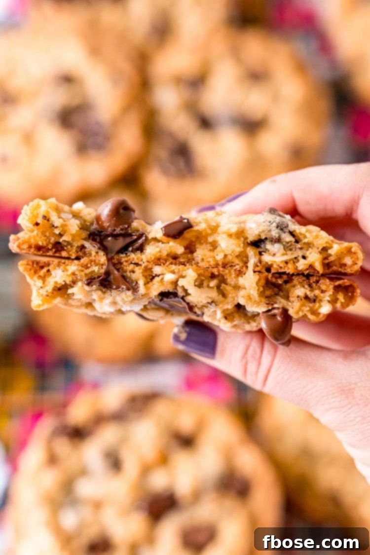 A woman's hand holding a broken in half ranger cookies to the camera so you can see the texture of the inside of the cookie.