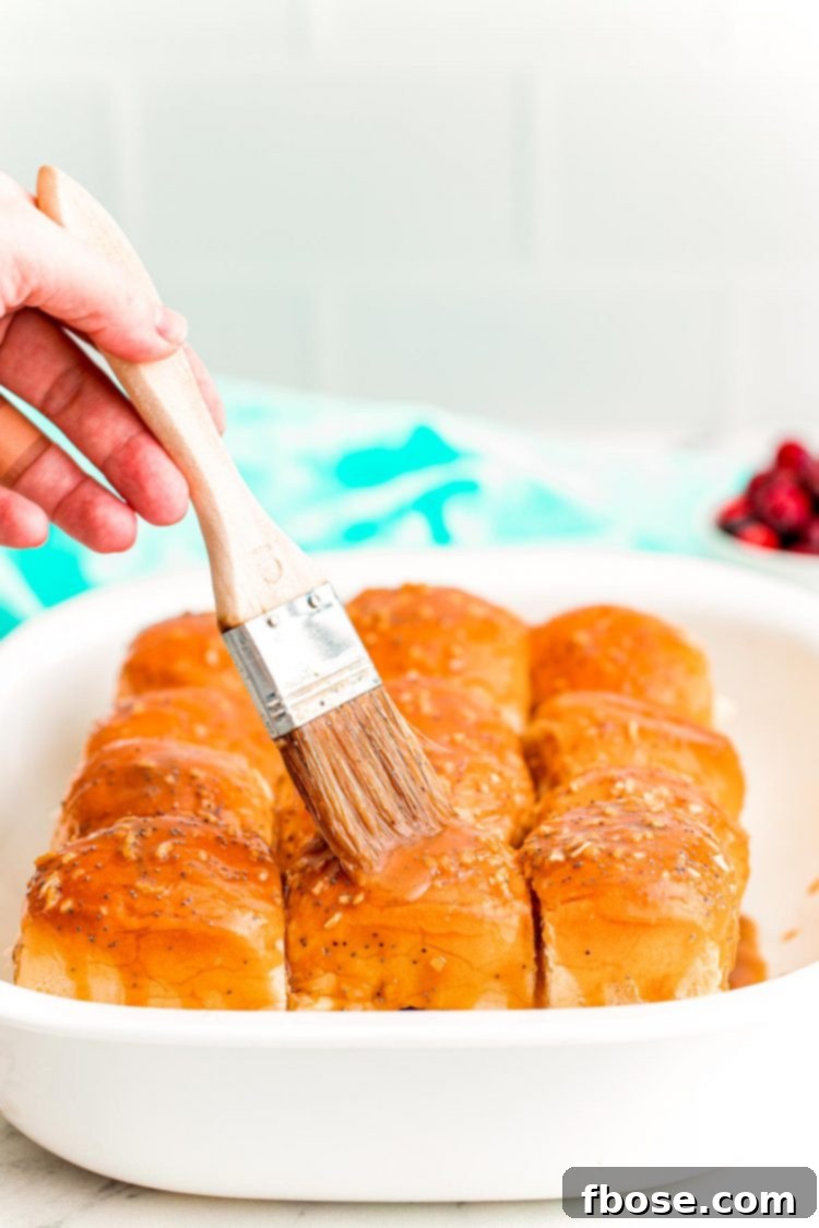 A woman's hand using a pastry brush to brush a butter mixture on top of the rolls of sliders in a white casserole dish.