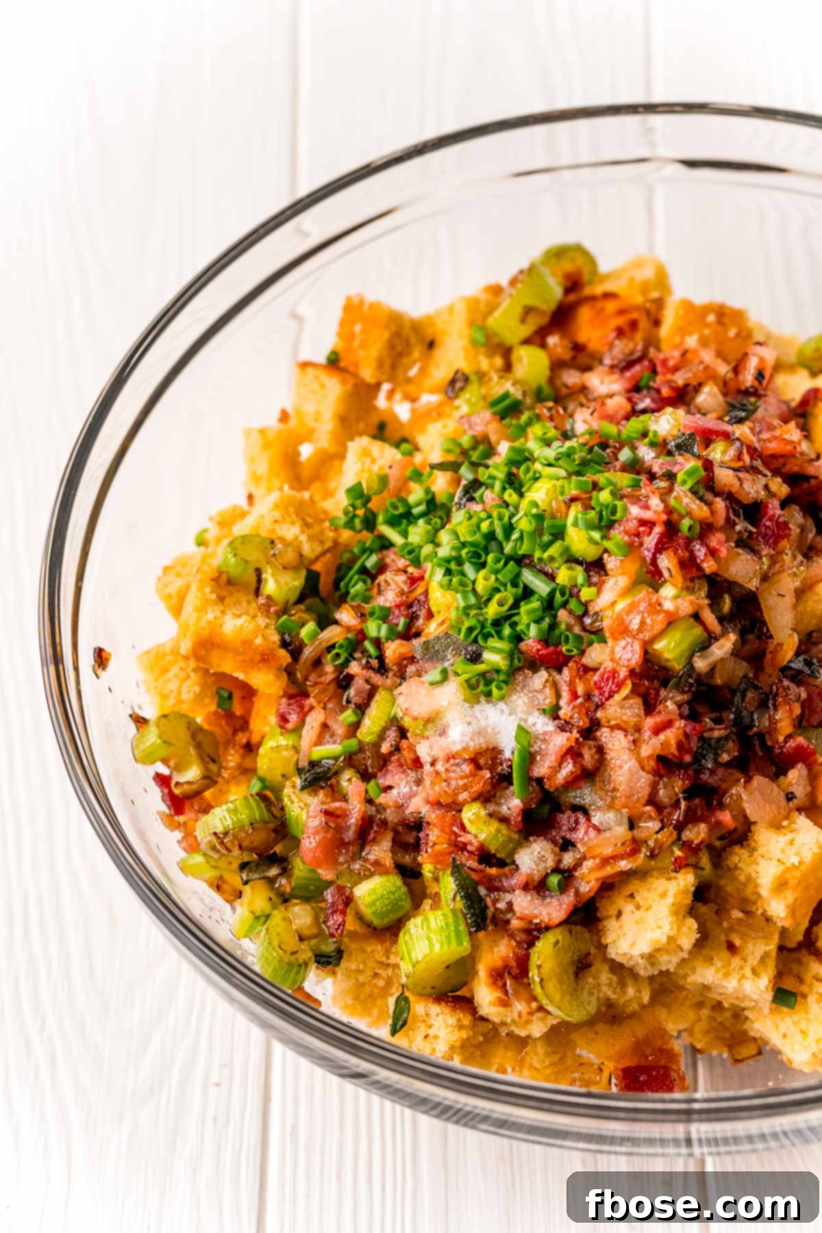 The Ultimate Cornbread Dressing 5 A large glass mixing bowl filled with cornbread cubes, sautéed celery, onions, chopped bacon, and fresh chives, ready for the liquid binders.