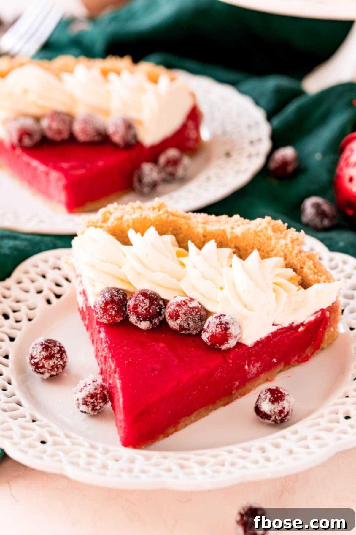 A slice of cranberry pie on a white plate topped with whipped cream and sugared cranberries. The pie features a golden shortbread crust and a vibrant red filling.