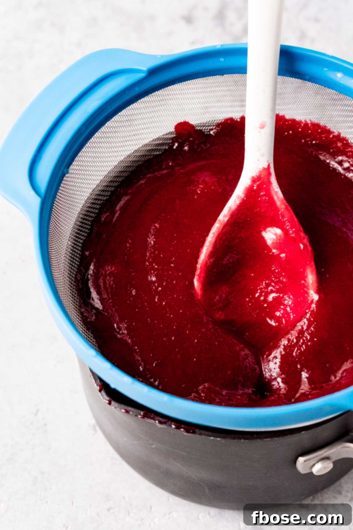 Smooth cranberry mixture being pressed through a fine-mesh strainer into a saucepan, with pulp collecting below.