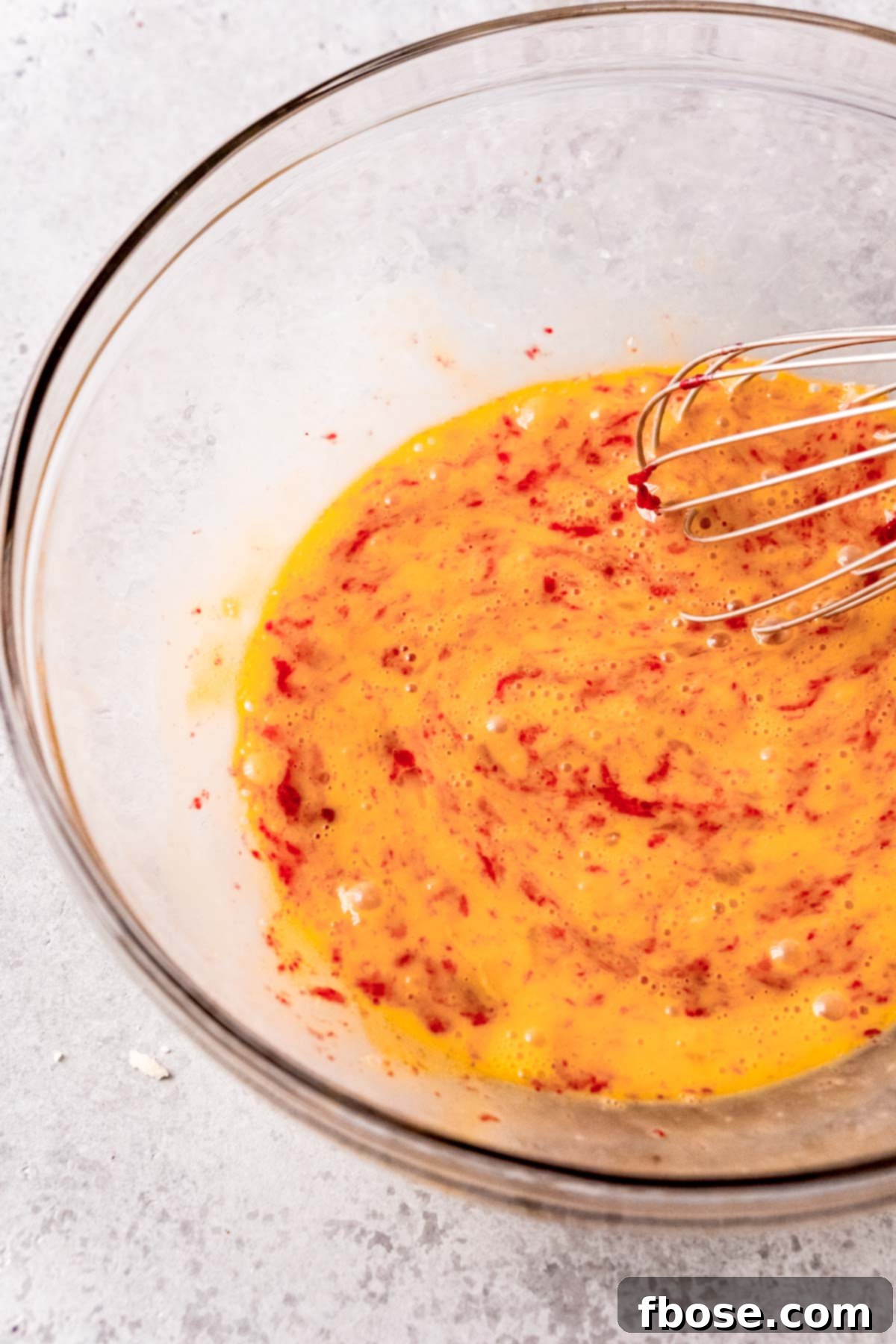 Beaten eggs in a clear glass mixing bowl, with a portion of the warm cranberry curd slowly mixed in for tempering.
