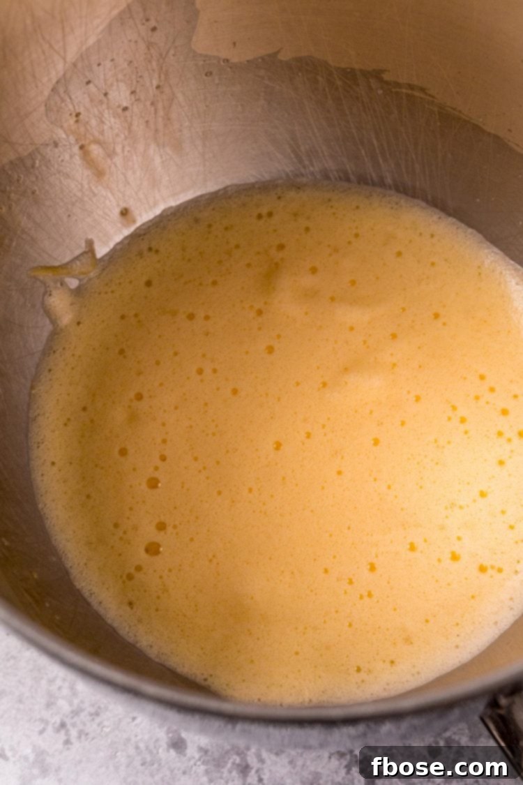A close-up view of light, foamy eggs being beaten in a stainless steel mixing bowl, showing their airy texture.
