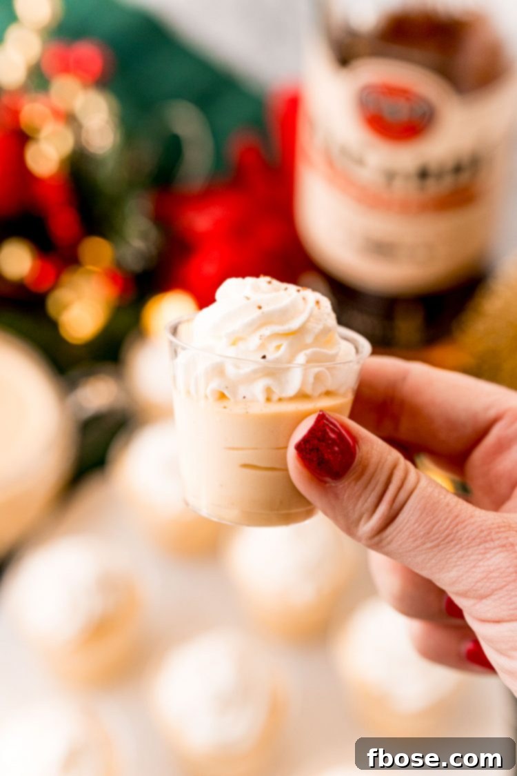A woman's hand holding a single eggnog pudding shot topped with whipped cream and a sprinkle of nutmeg.