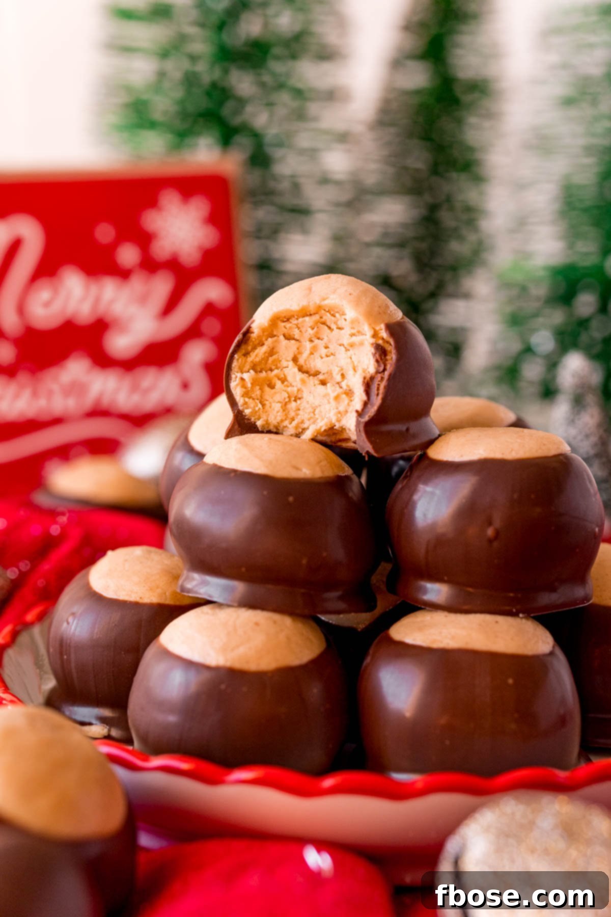 Close up photo of a plate of perfectly formed peanut butter buckeyes, with one featuring a bite taken out, set against a festive holiday background.